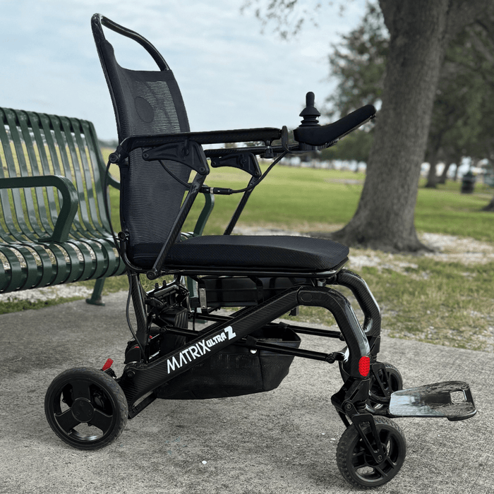Black Matrix 2 electric wheelchair on a sidewalk with a park bench and trees in the background.