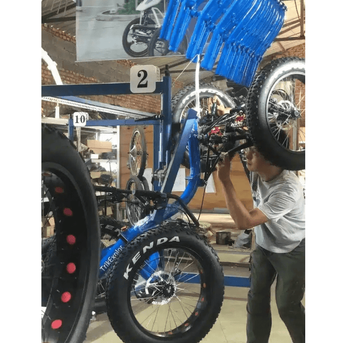 An indoor scene of a workshop with a worker adjusting a blue and black electric fat tire recumbent quad on a rack, surrounded by other trikes.