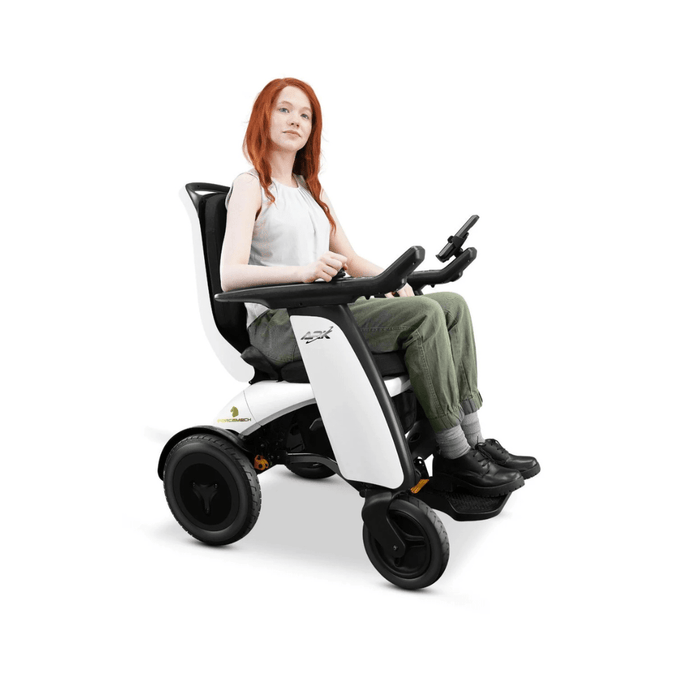 A person with long red hair sits in a modern Forcemech ARK All-Terrain Heavy Duty Folding Powerchair, equipped with Bluetooth speakers. They wear a white shirt, green pants, and black shoes. The sleek design features large rear wheels and a joystick control panel for enhanced mobility.