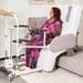 A woman on the starsleep arlo rotating bed in  a floral robe sits in a white and gray motorized bed with side rails, while another person at left holds the walker frame. The room has light neutral walls and a beige rug.